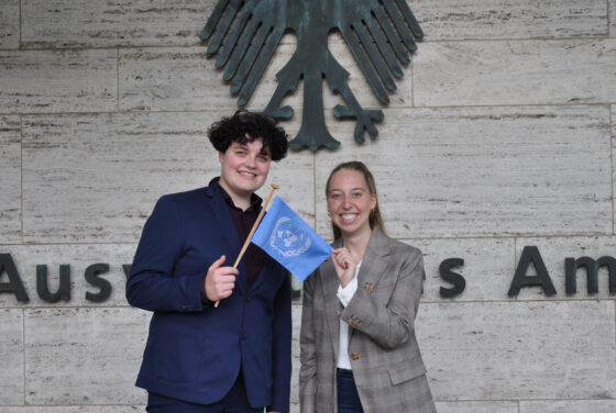 Lew und Ilka halten eine kleine UN-Flagge. Sie tragen Anzug und Blazer und stehen vor einer Mauer, auf der das Emblem des Auswäritgen Amts abgebildet ist.
